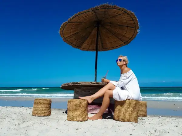 Kobieta w białej sukience siedzi pod parasolem na plaży na Djerbie. Uważać na słońce i upał.