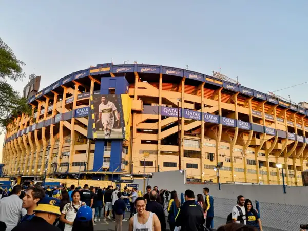 Tłum kibiców przed stadionem La Bombonera. Wielki baner z piłkarzem zdobi żółto-niebieską fasadę.