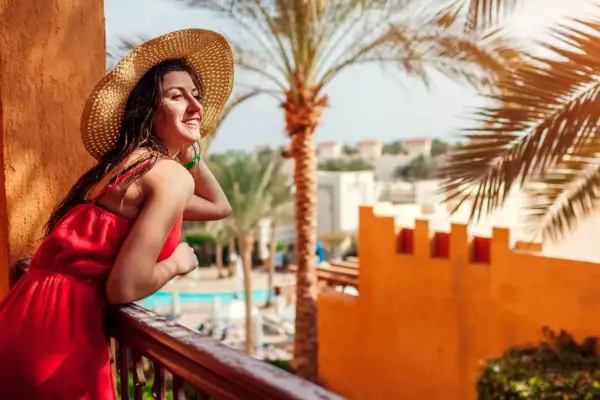 Uśmiechnięta kobieta w słomkowym kapeluszu i czerwonej sukience podziwia widok z balkonu. Idealna sharm el sheikh pogoda marzec.
