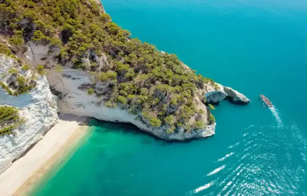Włochy regiony turystyczne: białe klify, piaszczysta plaża i turkusowe morze. Statek wycieczkowy płynie obok skalnego łuku.